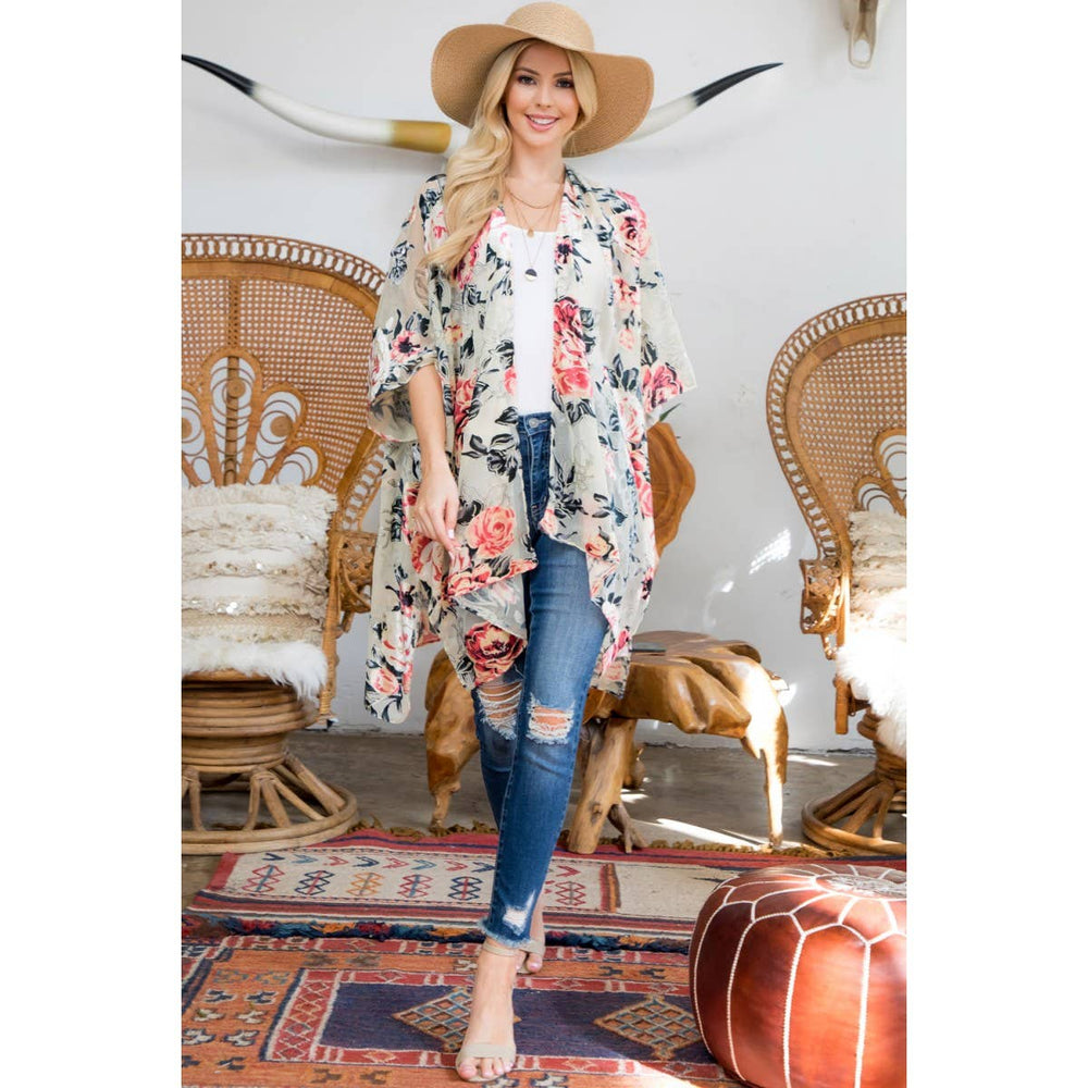 A woman stands in a boho-style room wearing a cream floral kimono cardigan over a white top and blue jeans, with a wide-brim tan hat and bare feet. Wicker chairs, patterned rugs, and a brown pouf are visible in the room.