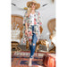 A woman stands in a boho-style room wearing a cream floral kimono cardigan over a white top and blue jeans, with a wide-brim tan hat and bare feet. Wicker chairs, patterned rugs, and a brown pouf are visible in the room.