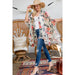 A woman models a bohemian-style floral kimono cardigan over a white top and blue jeans, standing in a styled room with rattan chairs and rugs.