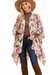 A woman modeling a long, open-front floral kimono cardigan with wide sleeves over a brown turtleneck, blue jeans, and brown knee-high boots, while wearing a wide-brimmed brown hat against a plain white background.