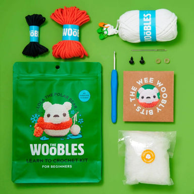 Theee Woothy Woolly Bits 'The Woobles' Learn to Crochet Kit for Beginners, laid out on a bright green background. The kit includes a green pouch with a polar bear illustration, a blue-handled crochet hook, white yarn, black yarn, red yarn, a brown card, white stuffing, and small metal findings.