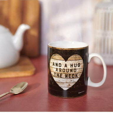Mug with a heart design and text on a wooden surface with a teapot and spoon in the background.