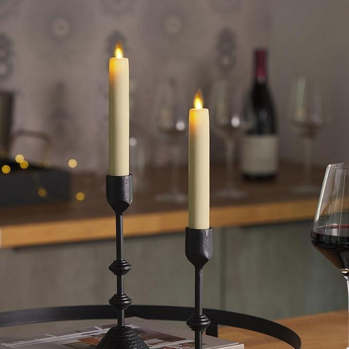 Two lit candles in black holders on a table with a blurred background of wine bottles and glasses.