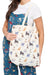 A woman wearing a white t-shirt and blue patterned pajama set holds a canvas tote bag. The tote bag is cream-colored with an all-over print of illustrated cats in various colors.