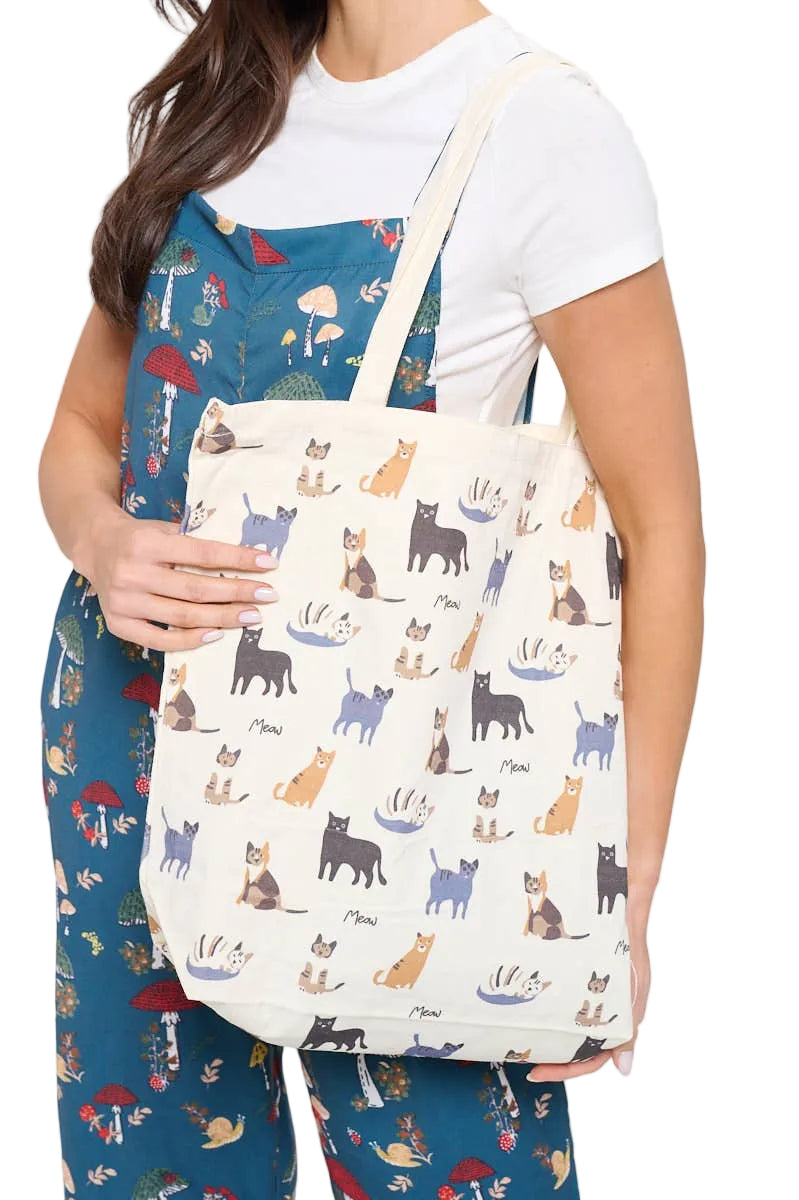 A woman wearing a white t-shirt and blue patterned pajama set holds a canvas tote bag. The tote bag is cream-colored with an all-over print of illustrated cats in various colors.
