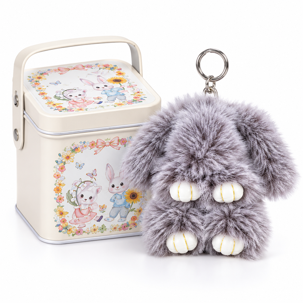 A gray plush bunny keychain with white paw pads and a metal key ring, shown next to a cream-colored metal tin with a floral bunny illustration on the lid and front.