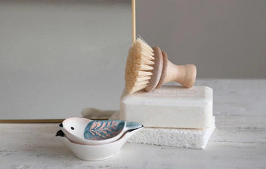 Bamboo dish brush, soap bar, and ceramic bird-shaped container on a light surface.