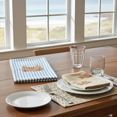 A styled dining table set near a window with a beach view. The table includes white plates, clear drinking glasses, a woven placemat, and two embroidered napkins: one blue and white striped with a crab motif, and one cream with a matching crab motif.