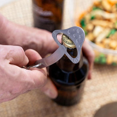 Person using a bottle opener on a beer bottle with food in the background
