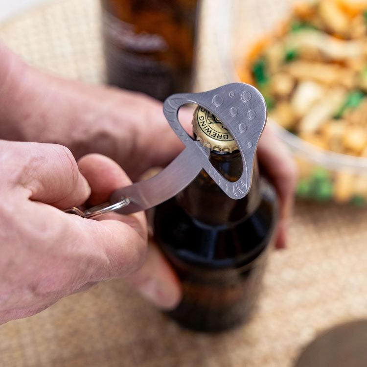 Person using a bottle opener on a beer bottle with food in the background