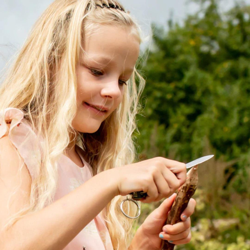 Young girl outdoors holding a small object with a knife, surrounded by greenery.