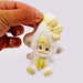 Small plush toy with blonde hair and a yellow outfit held by a hand on a white background