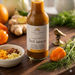 Bottle of Finch + Fennel hot sauce on a wooden surface with ingredients like oranges, ginger, and peppers.