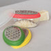 Small grater with colorful handles next to a container of grated cheese on a white surface.