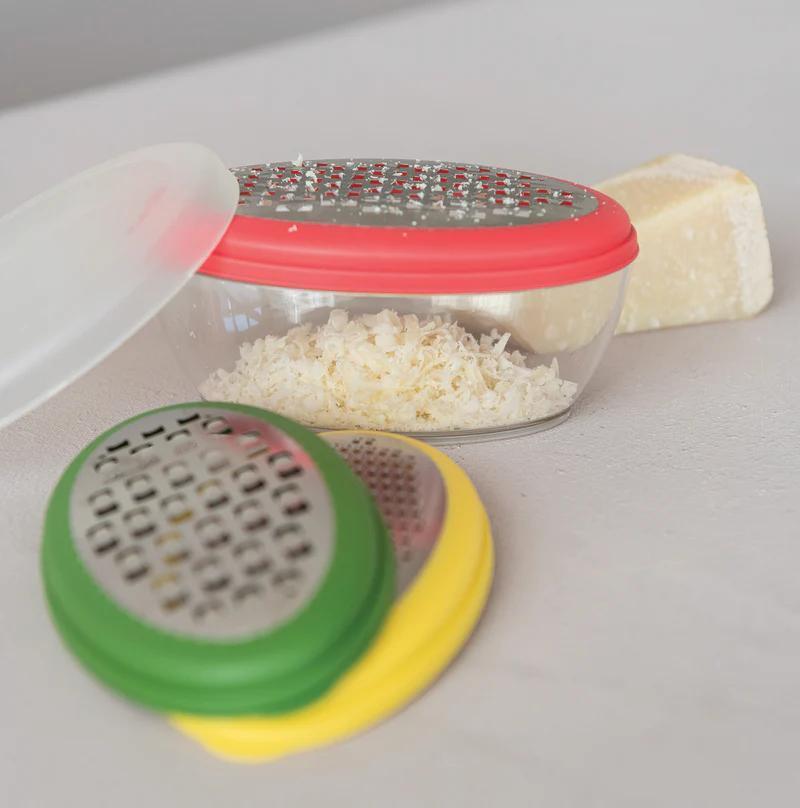 Small grater with colorful handles next to a container of grated cheese on a white surface.