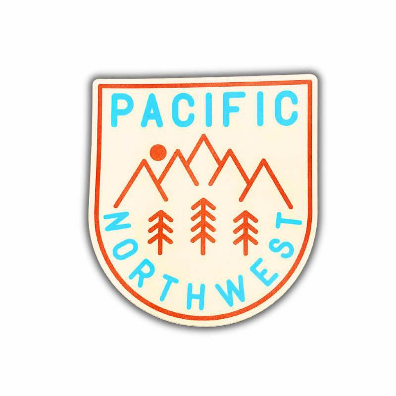 A shield-shaped sticker featuring the words 'PACIFIC NORTHWEST' in light blue, with orange mountain peaks, trees, and a sun on a cream background, shown on a white background.