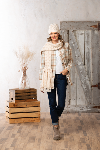Woman wearing a plaid jacket, scarf, and hat in a rustic setting with wooden crates and a wooden door.