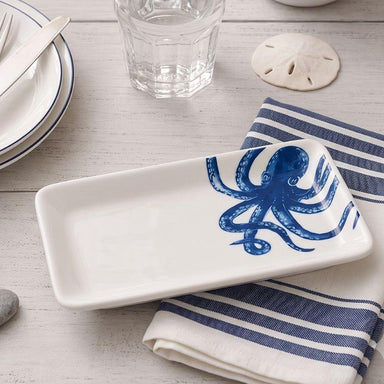 A white rectangular ceramic serving platter with a blue octopus design, styled on a striped napkin over a light wood tabletop.