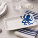 A white rectangular ceramic serving platter with a blue octopus design, styled on a striped napkin over a light wood tabletop.