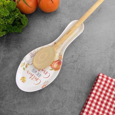 A white ceramic serving spoon with a floral design and the words 'Gettysburg Made with Love' resting on a gray stone surface, with a wooden spoon placed inside it. A red-and-white checkered cloth and greenery are partially visible in the corners.