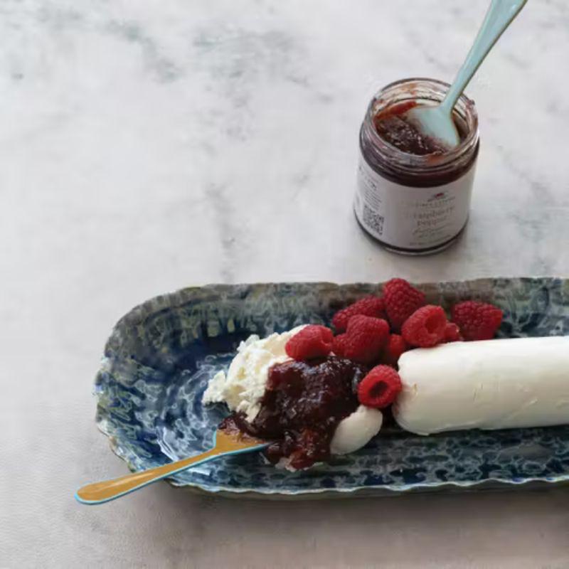 Jar of raspberry jam with a spoon, placed on a marble surface next to a dessert with whipped cream and raspberries.