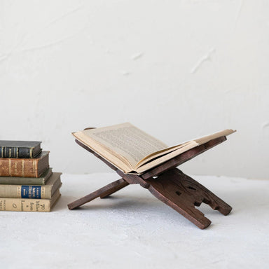Wooden book stand holding an open book with a stack of books on a white surface.