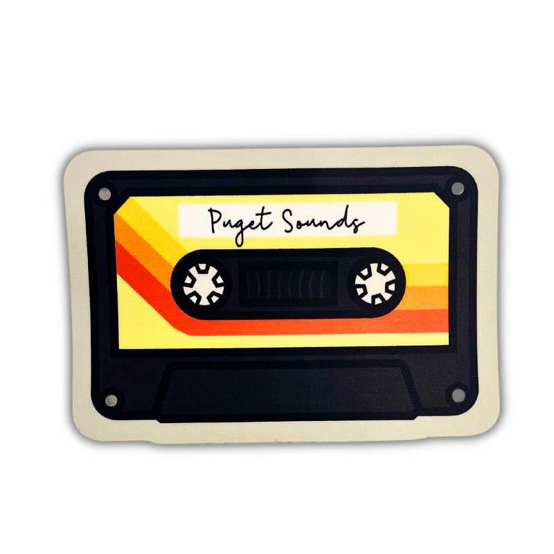 A retro cassette tape-shaped sticker or decal with a cream border, black inner frame, and a yellow label featuring orange stripes. The label reads 'Puget Sounds' in black script.