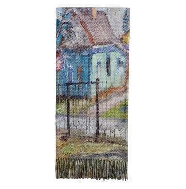 A rectangular woven textile with fringe along the bottom edge, featuring an artistic depiction of a house and fence in soft blue, green, brown, and gray tones, shown against a light gray background.