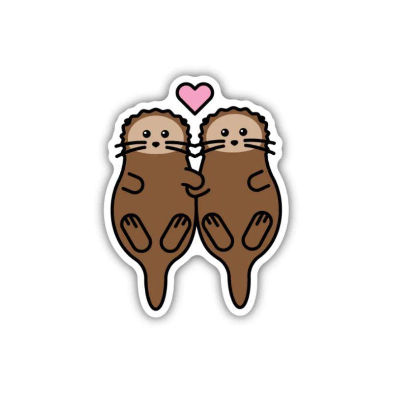 A die-cut sticker featuring two cute cartoon brown otters facing each other with a small pink heart above them, shown on a plain white background.
