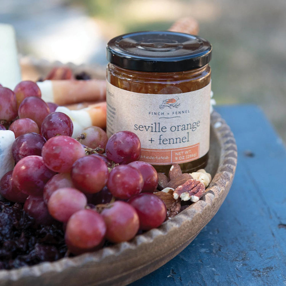Jar of 'Seville Orange + Fennel' jam on a plate with grapes and nuts outdoors