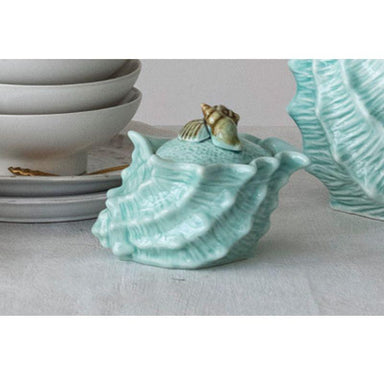 Turquoise ceramic shell-shaped container with a small shell on top, placed on a light surface.