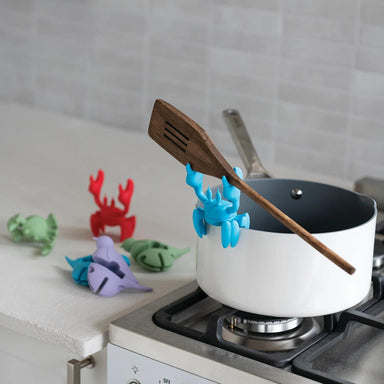 White pot on a stove with a wooden spoon, surrounded by colorful animal-shaped kitchen tools.