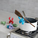 White pot on a stove with a wooden spoon, surrounded by colorful animal-shaped kitchen tools.