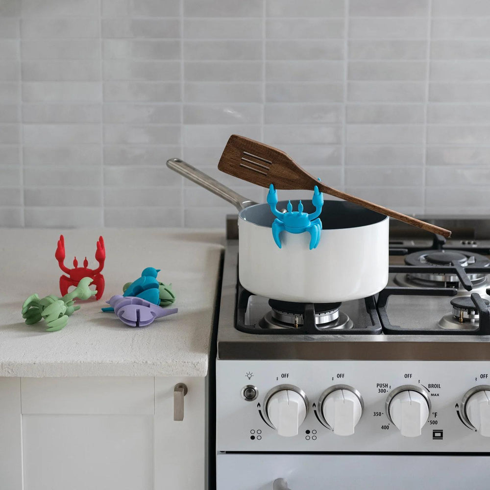 A kitchen scene with a white saucepan on a gas stove, a wooden spatula resting on the handle, and a bright blue crab-shaped pot holder clipped to the pot rim. Small colorful crab-shaped pot holders are also placed on the countertop against a light tile backsplash.