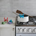 A kitchen scene with a white saucepan on a gas stove, a wooden spatula resting on the handle, and a bright blue crab-shaped pot holder clipped to the pot rim. Small colorful crab-shaped pot holders are also placed on the countertop against a light tile backsplash.