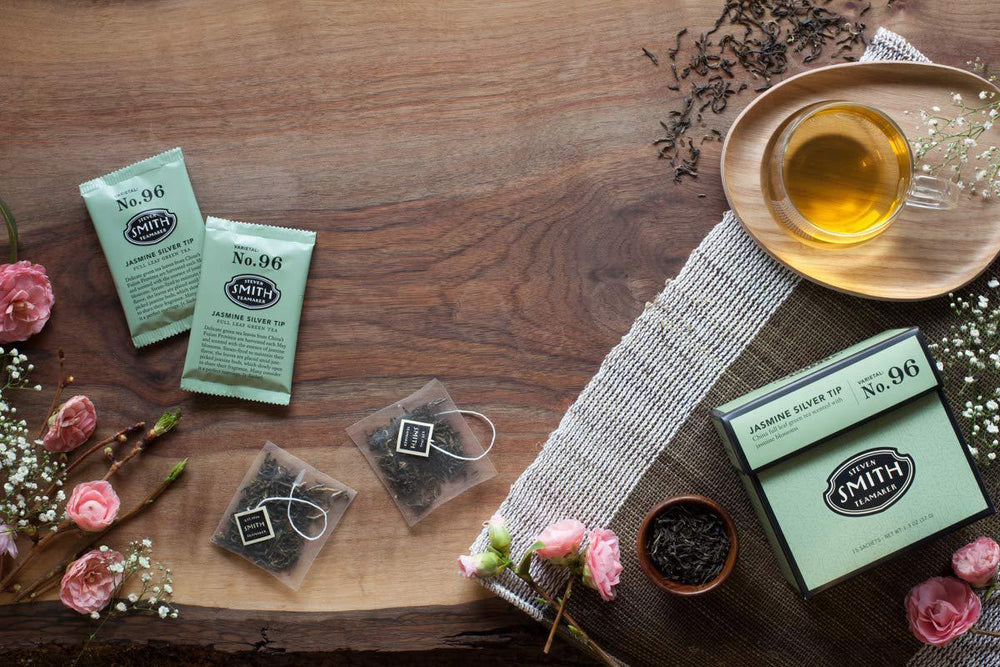 Tea packaging and loose tea on a wooden surface with flowers and a teacup.