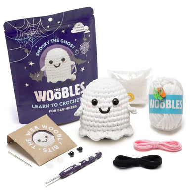 The We Woobles 'Snooky the Ghost' beginner crochet kit arranged on a white background, including a purple branded pouch, a crocheted ghost figure, white yarn, pink and black thread, metal tools, and a brown paper bag.