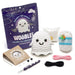 The We Woobles 'Snooky the Ghost' beginner crochet kit arranged on a white background, including a purple branded pouch, a crocheted ghost figure, white yarn, pink and black thread, metal tools, and a brown paper bag.