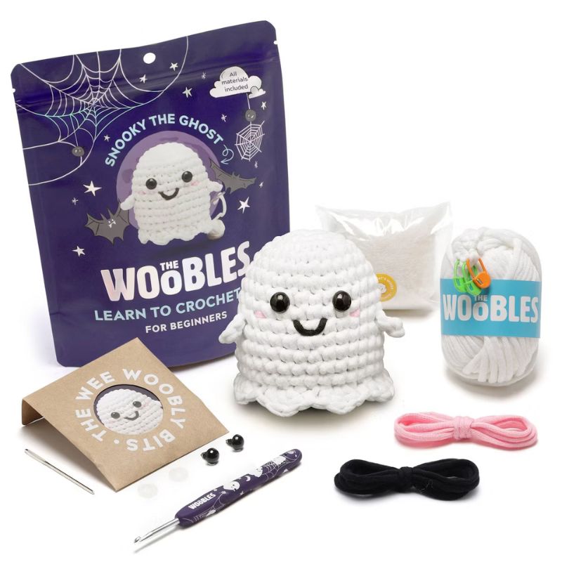 The We Woobles 'Snooky the Ghost' beginner crochet kit arranged on a white background, including a purple branded pouch, a crocheted ghost figure, white yarn, pink and black thread, metal tools, and a brown paper bag.
