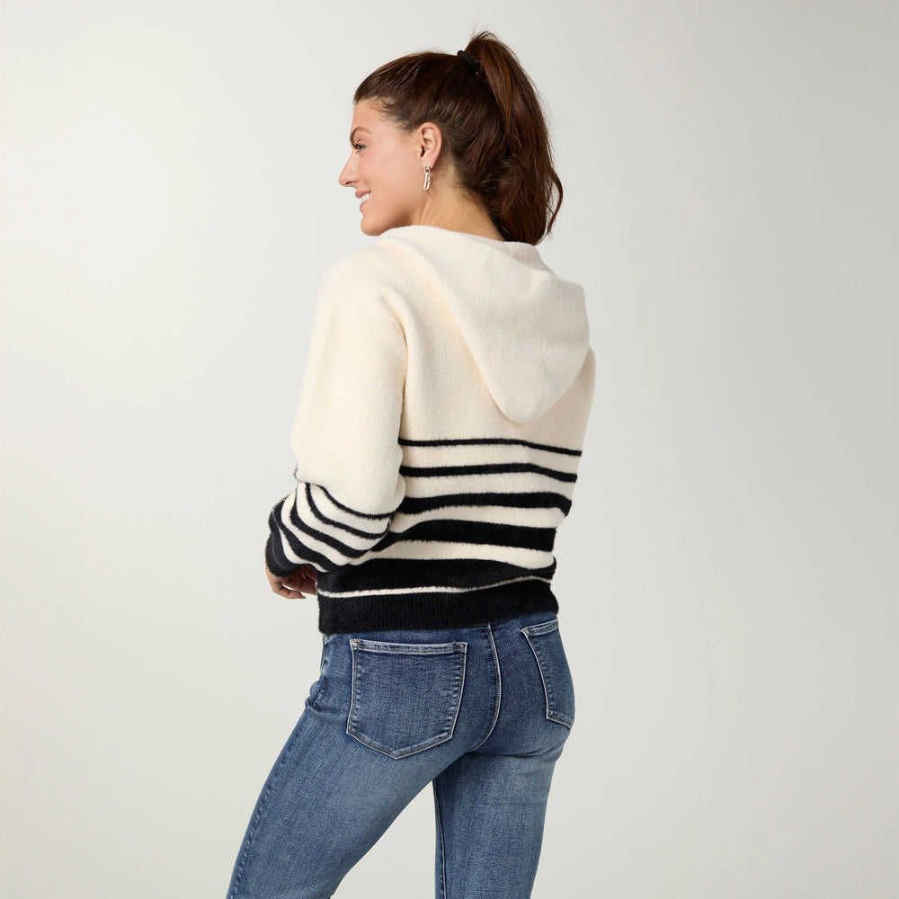 A woman shown from the back wearing a cream knit hoodie with black horizontal stripes on the sleeves and lower back, paired with blue jeans, against a light gray studio background.