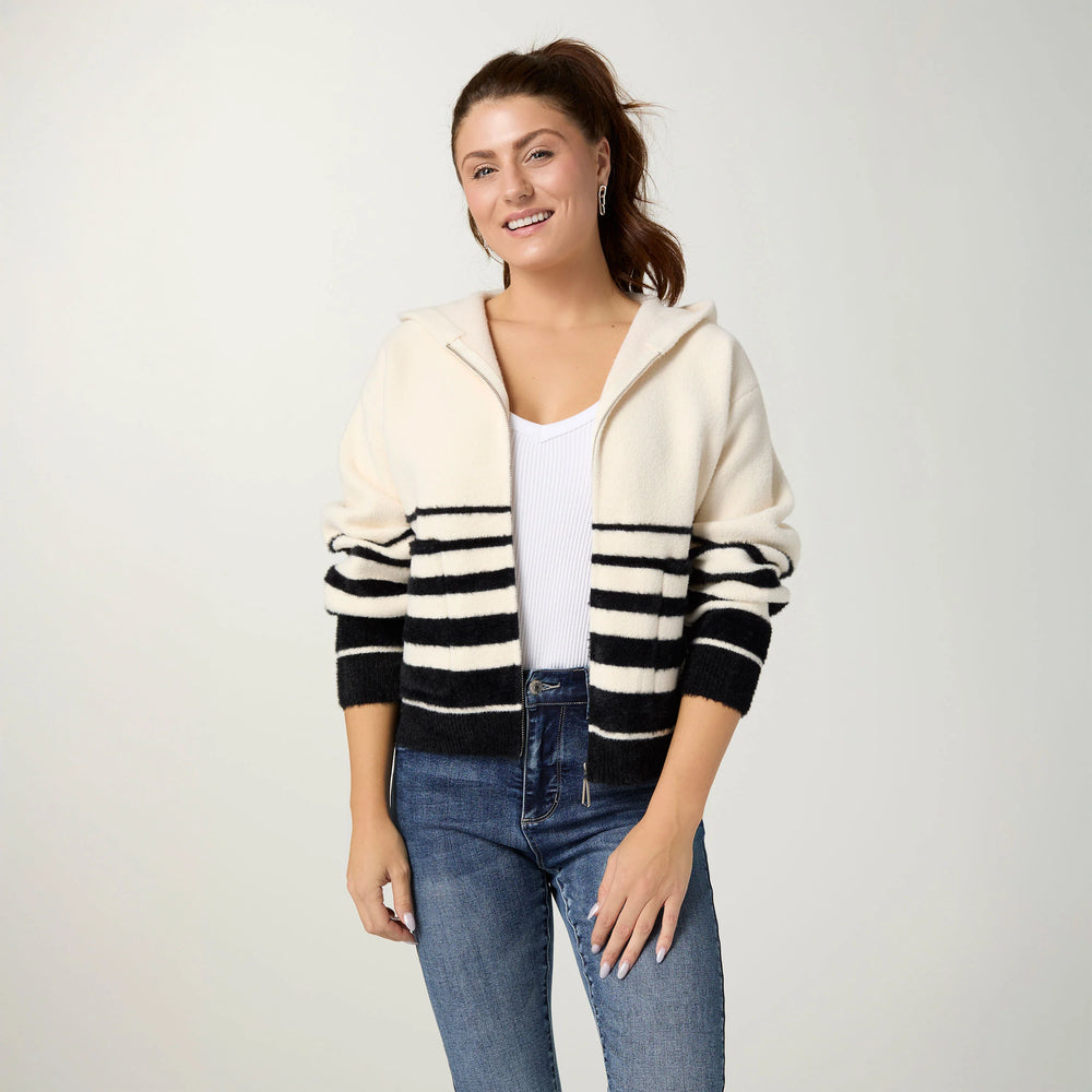 A woman modeling a cream and black striped knit zip-up hoodie with a hood, worn over a white tank top and blue jeans, against a light gray studio background.