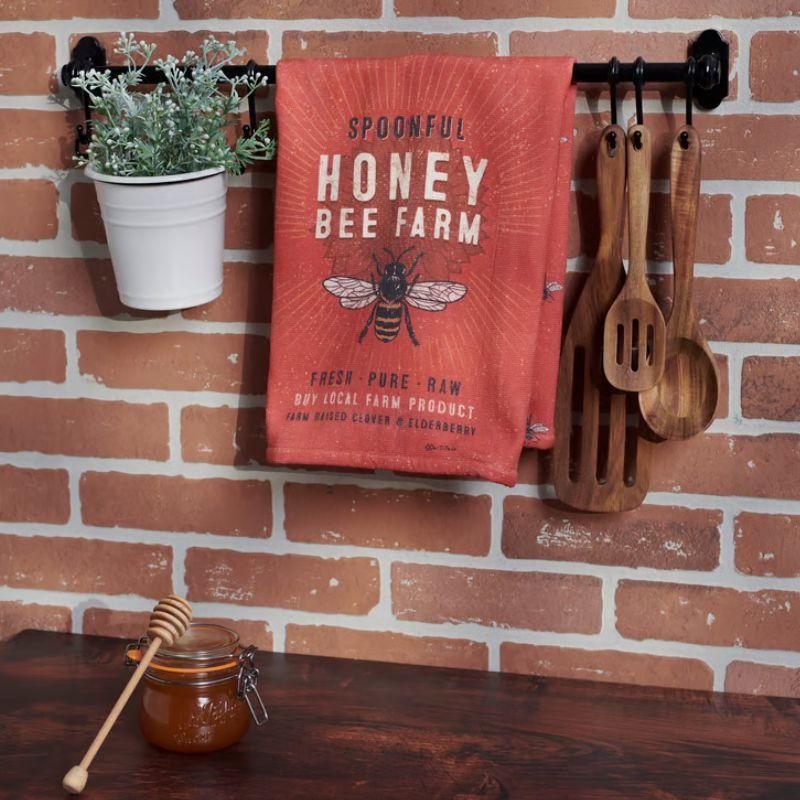 Red towel with 'Spoonful Honey Bee Farm' branding hanging on a brick wall with wooden utensils and a jar of honey below.