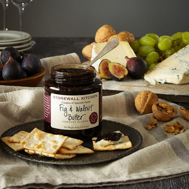 Stonewall Kitchen Fig & Walnut Butter jar on a plate with crackers and figs, surrounded by cheese and grapes.