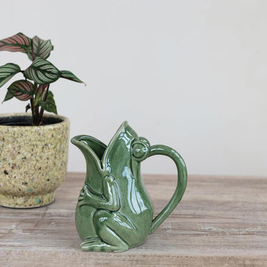 Green ceramic frog-shaped pitcher on a wooden surface with a plant in the background