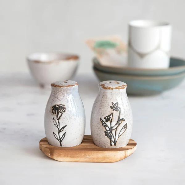 Two ceramic salt and pepper shakers with floral designs on a wooden coaster, with bowls in the background.