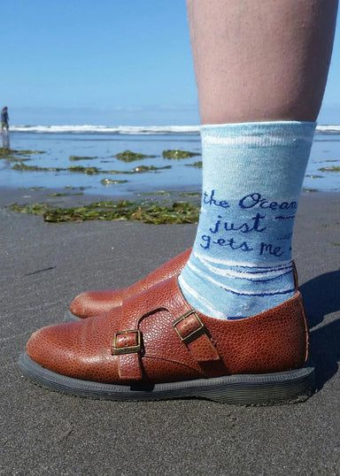 Person wearing brown shoes with a sock featuring ocean-themed text on a beach.