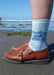 Person wearing brown shoes with a sock featuring ocean-themed text on a beach.