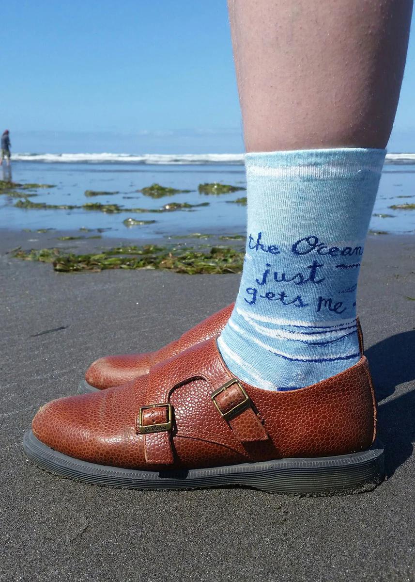 Person wearing brown shoes with a sock featuring ocean-themed text on a beach.