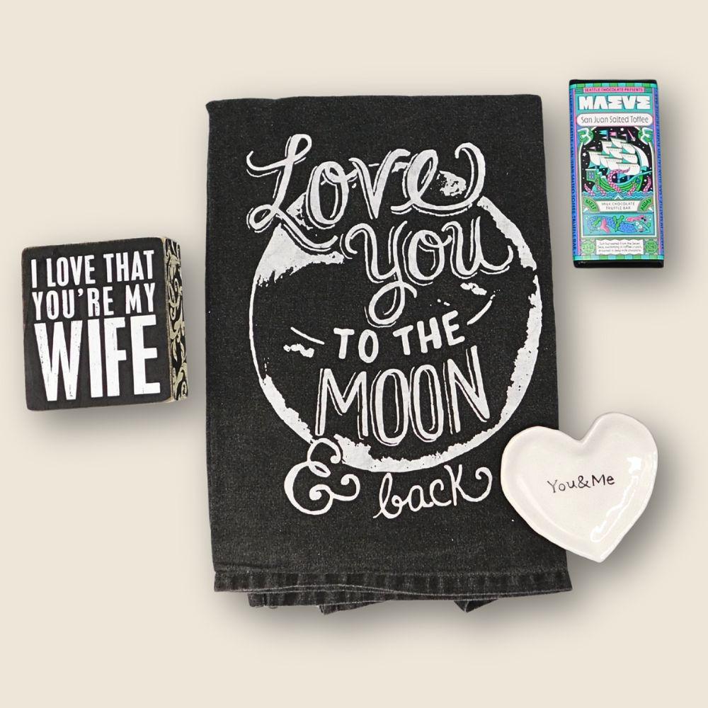 Black towel with 'Love you to the moon & back' text, 'I love that you're my wife' card, and heart-shaped dish on a light background.
