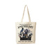 An off-white canvas tote bag with a gothic apothecary illustration, hanging on a gray wall next to a blue denim jacket. The bag features the text 'Handcrafted Feathered Potions', 'APOTHECARY', and 'WHERE MAGIC MEETS MIDNIGHT' with an illustration of a raven, glass bottles, and flowers.